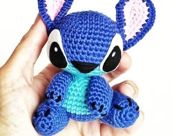 May include: A blue and turquoise crocheted Stitch plush toy with pink ears and black eyes. The toy is sitting in a person's hand.