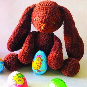 Puede incluir: Un conejito de peluche de color marrón tejido a crochet sentado con huevos de Pascua coloridos. Los huevos están envueltos en papel de aluminio y tienen un diseño de pollito.