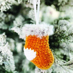 Puede incluir: Una pequeña media de punto de ganchillo naranja con un diseño de copo de nieve blanco de ganchillo colgando de un árbol de Navidad nevado.