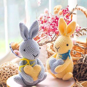 May include: Two crocheted bunny rabbits holding heart-shaped crocheted objects. The bunny on the left is light blue with a yellow heart and a yellow ribbon. The bunny on the right is yellow with a blue heart and a blue ribbon. The bunnies are sitting on a heart-shaped box with two yellow and one purple crocheted eggs.