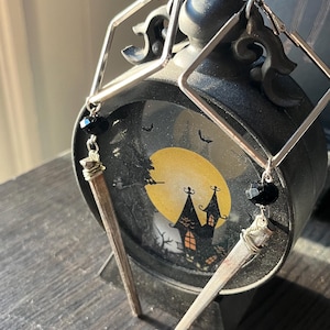 Puede incluir: Pendientes de aro cuadrados plateados con una piedra preciosa negra y un dije en forma de clavo. Los pendientes se exhiben frente a un reloj decorativo negro con una imagen de Halloween de una casa embrujada y una luna llena.
