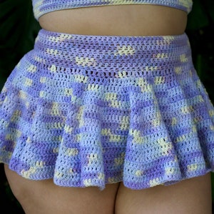 Puede incluir: Falda de ganchillo en tonos lavanda, azul claro y amarillo pálido. La falda tiene una silueta acampanada, estilo patinador. La prenda hecha a mano es parte de un conjunto a juego, con un vistazo de un top visible encima de la falda.