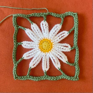 Op de afbeelding: Een gehaakt vierkant met een witte madeliefje in het midden. De madeliefje heeft een gele kern en groene bladeren. Het vierkant is gemaakt van groen garen.