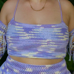 Puede incluir: Un conjunto de top y falda de crochet de color lavanda y blanco con un detalle de lazada en los hombros. El top es de estilo corto y la falda es de estilo de cintura alta.
