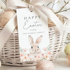 Może przedstawiać: Biały wiklinowy kosz z kremową wstążką i etykietą z napisem "Happy Easter" z ilustracją królika. Etykieta jest spersonalizowana z "The Harris Family". Kosz zawiera dekoracyjne jajka.