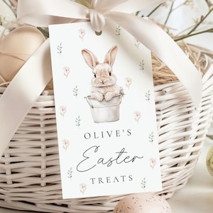 May include: A white Easter gift tag with a watercolor illustration of a bunny in a bucket, surrounded by floral accents. The tag reads "Olive's Easter Treats" in elegant script. A white basket and ribbon are in the background.
