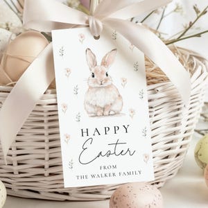 May include: A white Easter gift tag with a watercolor illustration of a bunny and floral accents. The tag reads "Happy Easter" and "From The Walker Family". The tag is attached to a white wicker basket with a cream-colored ribbon.