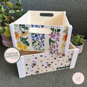 May include: Two stacked wooden crates with cut-out handles. The top crate features a floral pattern with yellow, purple, and blue flowers and a butterfly. The bottom crate displays a pattern of colourful polka dots and bees with the text Emma Bridgewater.