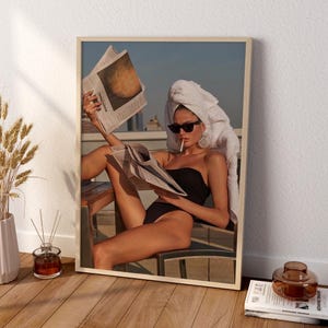 May include: Framed art print featuring a woman in a black swimsuit, sunglasses, and a white towel on her head, reading a newspaper. The print has a neutral wood frame and is displayed on a white wall. The image evokes a sense of leisure and relaxation.