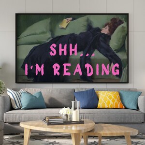 Op de afbeelding: Een ingelijste print van een schilderij van een vrouw die op een bank ligt en een boek leest. De tekst "SHH... I'M READING" staat in roze letters over de afbeelding.