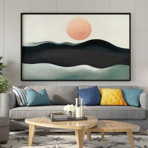 May include: A framed abstract painting with a large, peach-colored circle in the center, representing the sun, above a series of dark blue and green hills. The painting is hanging on a white wall above a gray sofa with blue and yellow throw pillows.