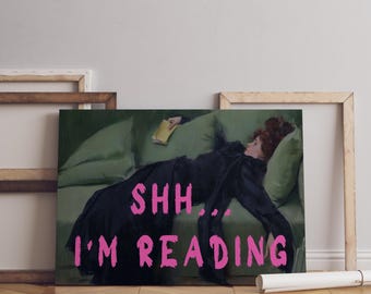 May include: A rectangular wall art print featuring a painting of a person in a black dress reclining on a green sofa while holding a book. The text SHH... I'M READING is overlaid in bright pink, bold, sans-serif capital letters across the center.