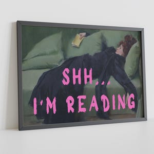 Op de afbeelding: Een ingelijste print van een schilderij van een vrouw die op een groene bank ligt en een boek leest. De tekst "SHH... I'M READING" staat in roze letters onderaan de afbeelding.