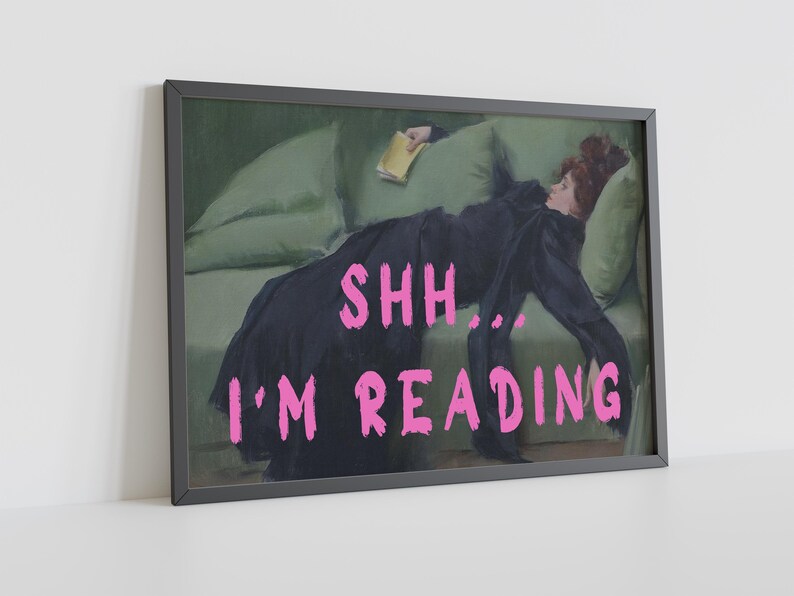 Op de afbeelding: Een ingelijste print van een schilderij van een vrouw die op een groene bank ligt en een boek leest. De tekst "SHH... I'M READING" staat in roze letters onderaan de afbeelding.