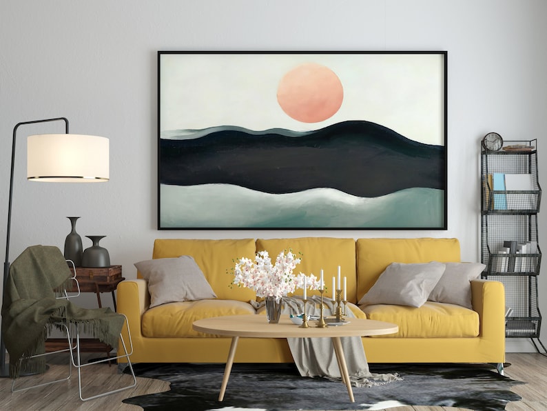 May include: A yellow sofa with two pillows and a throw blanket sits in front of a large abstract painting of a pink sun setting over a dark blue and green landscape. A coffee table with a vase of white flowers sits in front of the sofa.