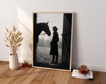 May include: A framed black and white wall art print featuring a silhouette of a person standing in a barn doorway facing a horse. The scene depicts a field in the background. The print is displayed in a light wood frame on a wooden floor.