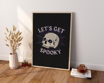 May include: A rectangular framed wall art print featuring a black background with a central illustration of a human skull resting on a purple spider web. The text LET'S GET SPOOKY is printed in white capital letters above and below the skull.