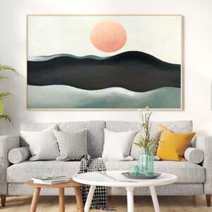 May include: Abstract painting of a landscape with a large pink sun in the sky and green and black hills. The painting is framed and hanging on a white wall above a gray sofa.