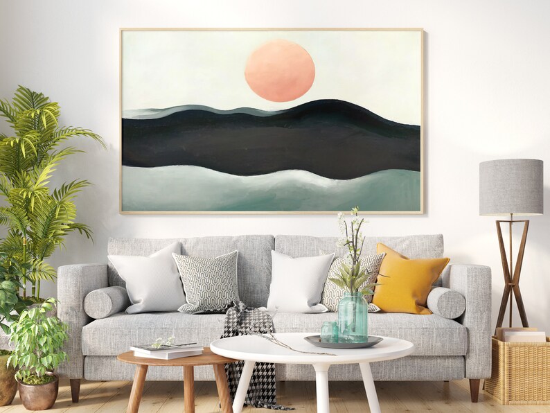 May include: Abstract painting of a landscape with a large pink sun in the sky and green and black hills. The painting is framed and hanging on a white wall above a gray sofa.