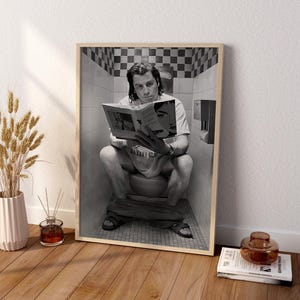 Pode incluir: Uma impressão de arte de parede emoldurada em preto e branco apresentando uma pessoa sentada num vaso sanitário a ler um livro. A pessoa veste uma t-shirt com o texto 'BANANA SLUG' e sandálias. O fundo mostra uma casa de banho azulejada.