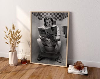 May include: A framed black and white wall art print featuring a person sitting on a toilet reading a book. The person wears a t-shirt with the text 'BANANA SLUG' and sandals. The background shows a tiled bathroom with a checkered border.