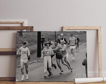 May include: A black and white photograph depicting a group of runners on a paved path. The participants wear athletic shirts and shorts with visible race bib numbers including 225, 261, and 295. The runners are in motion outdoors.
