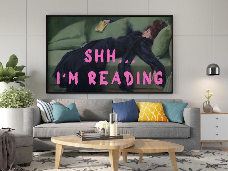 Op de afbeelding: Een ingelijste print van een schilderij van een vrouw die op een bank ligt en een boek leest. De tekst "SHH... I'M READING" staat in roze letters over de afbeelding.