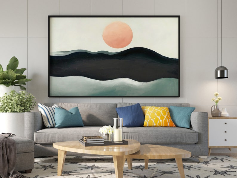 May include: A framed abstract painting with a large, peach-colored circle in the center, representing the sun, above a series of dark blue and green hills. The painting is hanging on a white wall above a gray sofa with blue and yellow throw pillows.