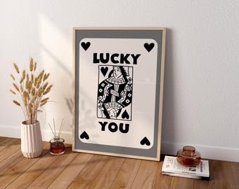 May include: A rectangular framed wall art print featuring a stylized playing card design with a white background, black border, and four black heart symbols in the corners. The text LUCKY YOU is printed in bold black letters above and below a central illustration of a queen figure.