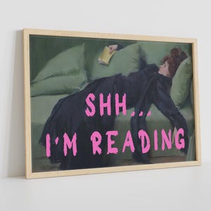 Op de afbeelding: Een ingelijste print van een schilderij dat een vrouw toont die op een bank ligt en een boek leest. De tekst "SHH... I'M READING" staat in roze letters onderaan de afbeelding.