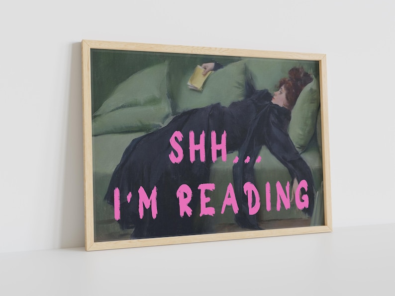 Op de afbeelding: Een ingelijste print van een schilderij dat een vrouw toont die op een bank ligt en een boek leest. De tekst "SHH... I'M READING" staat in roze letters onderaan de afbeelding.