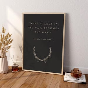 May include: A rectangular framed wall art piece featuring a dark charcoal background with the white text "WHAT STANDS IN THE WAY, BECOMES THE WAY." and "-MARCUS AURELIUS" above a white line drawing of a laurel wreath centered at the bottom.