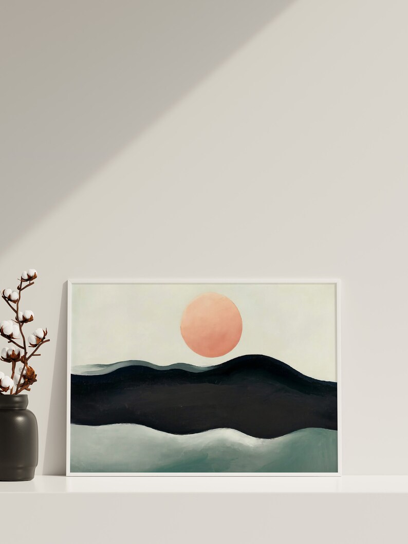May include: A framed abstract painting with a large, peach-colored circle in the center, representing the sun, above a series of dark blue and green hills.