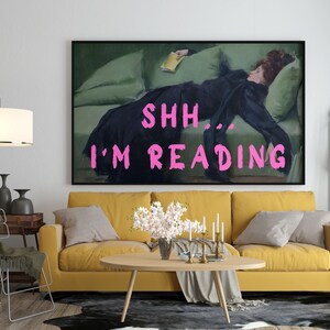 Op de afbeelding: Een gele bank met een schilderij van een vrouw die op een groene bank ligt. Het schilderij heeft de tekst "SHH... I'M READING" in roze letters.