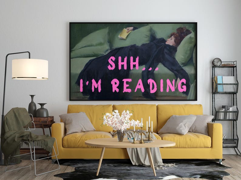 Op de afbeelding: Een gele bank met een schilderij van een vrouw die op een groene bank ligt. Het schilderij heeft de tekst "SHH... I'M READING" in roze letters.