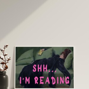 Op de afbeelding: Een ingelijste print van een schilderij van een vrouw die op een groene bank ligt en een boek leest. De tekst "SHH... I'M READING" staat in roze letters op de print.