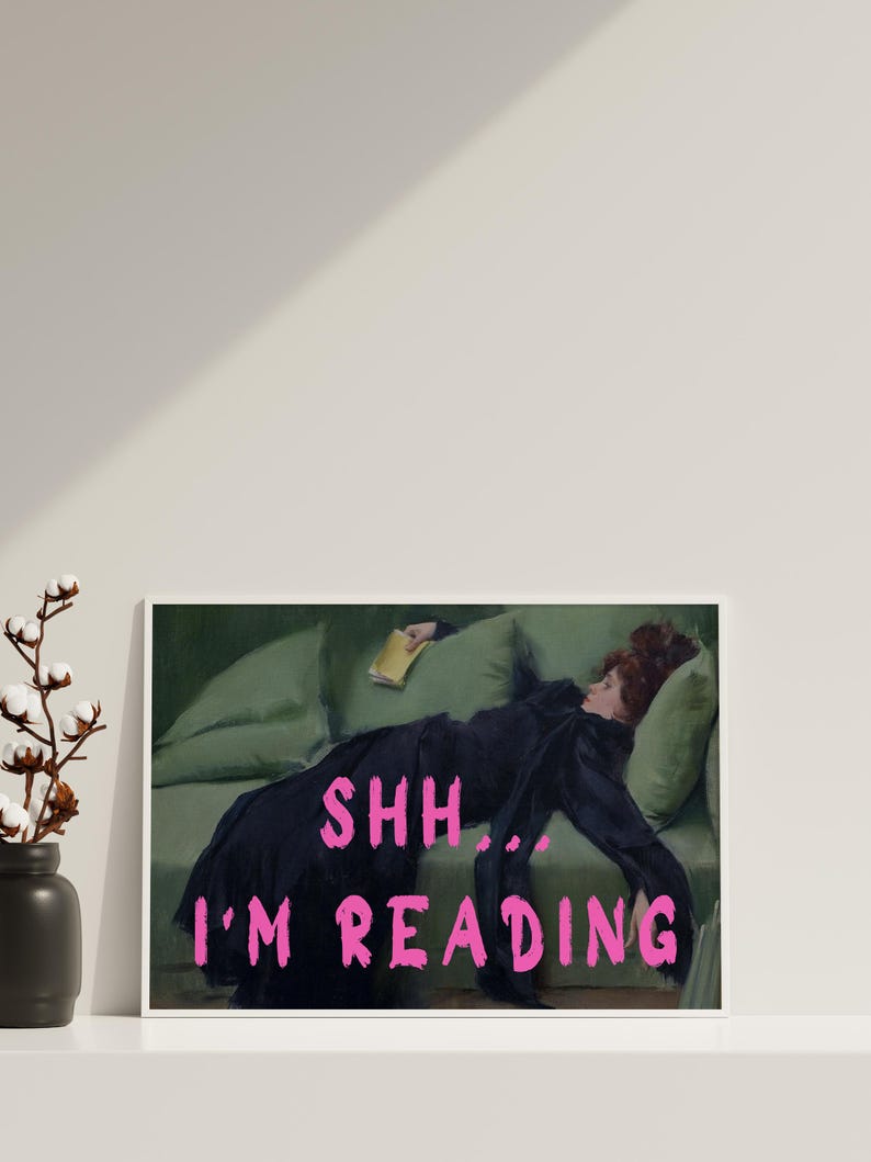 Op de afbeelding: Een ingelijste print van een schilderij van een vrouw die op een groene bank ligt en een boek leest. De tekst "SHH... I'M READING" staat in roze letters op de print.