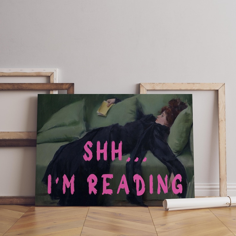 Op de afbeelding: Een schilderij van een vrouw die op een groene bank ligt en een boek leest. De tekst "SHH... I'M READING" staat in roze letters onderaan het schilderij.