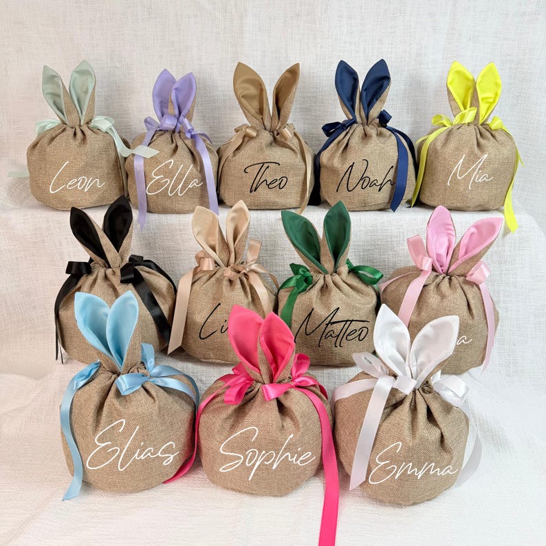 May include: A collection of burlap gift bags, each adorned with bunny ears in various colors like blue, pink, and yellow. Each bag is tied with a matching ribbon and features a name in elegant script.