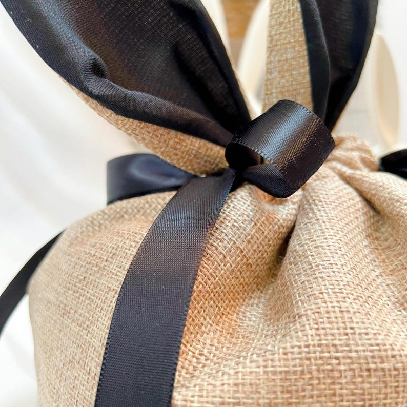May include: Close-up of a burlap gift bag tied with a black satin ribbon. The bag is made of a coarse, woven fabric in a natural tan color. The black ribbon is wrapped around the bag and tied in a bow at the top.