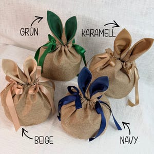May include: Four burlap gift bags with bunny ear accents. Each bag is a different color: green, caramel, beige, and navy. The bags are tied with matching ribbons and are suitable for Easter or spring gifting.