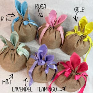 May include: Six burlap Easter gift bags with bunny ears and ribbon. Colors include blue, pink, yellow, mint, lavender, and flamingo. Text labels in German identify each color: Blau, Rosa, Gelb, Mint, Lavendel, and Flamingo.