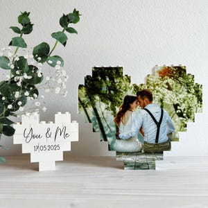 Op de afbeelding: Twee witte hartvormige fotolijsten gemaakt van in elkaar grijpende blokken. Eén lijst toont de tekst "You & Me" en de datum "17.05.2025". De andere lijst bevat een foto van een stel. Groene bladeren zijn zichtbaar.