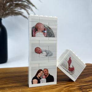 Peut inclure: Un pr&eacute;sentoir photo en briques Lego blanches avec trois images d'un nouveau-n&eacute; et un portrait de famille. Les images sont imprim&eacute;es sur les briques Lego, cr&eacute;ant un souvenir unique et personnalis&eacute;. Le pr&eacute;sentoir est sur une surface en bois.