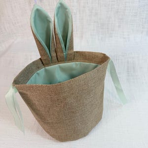 May include: A brown burlap Easter basket with light green accents. The basket has two upright bunny ears and a light green interior. Light green ribbons are attached to the sides of the basket.