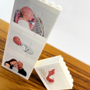 Peut inclure: Blocs blancs de style Lego avec des photographies imprim&eacute;es. Un bloc affiche trois images : un b&eacute;b&eacute; endormi, un autre b&eacute;b&eacute; endormi et un couple tenant un b&eacute;b&eacute;. Un deuxi&egrave;me bloc montre les pieds d'un b&eacute;b&eacute;. Les blocs sont sur une surface en bois.