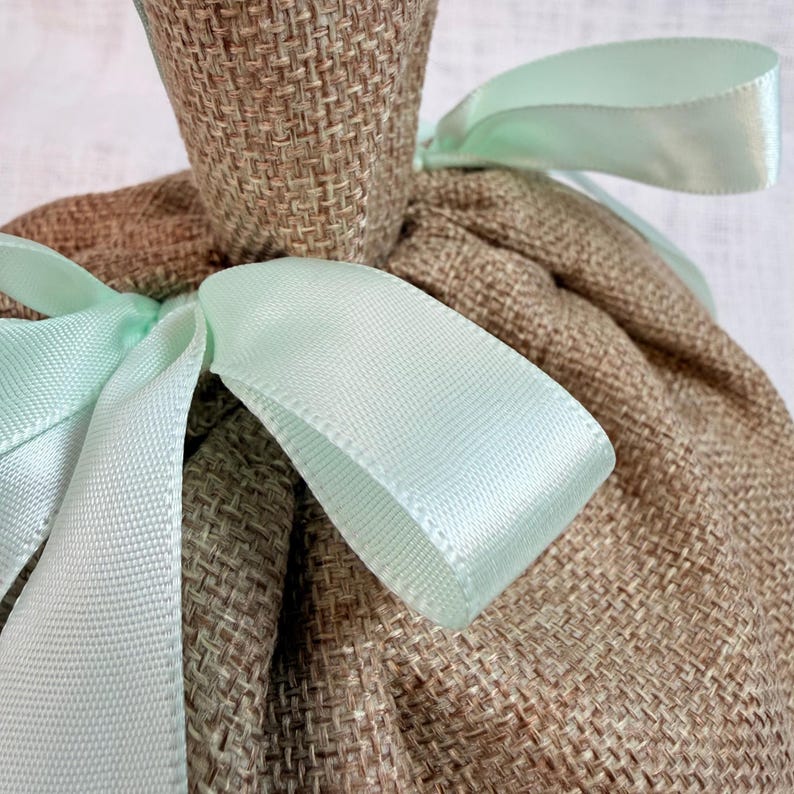 May include: Close-up of a burlap gift bag tied with a light green satin ribbon. The bag is a natural brown color, and the ribbon adds a delicate touch. The texture of the burlap is visible.