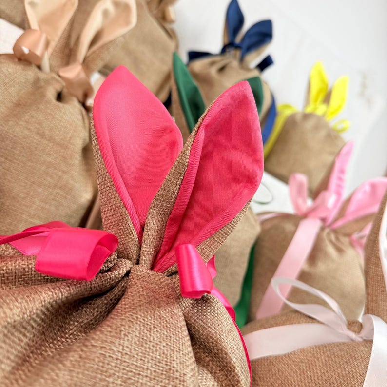 May include: Close-up of several burlap gift bags with bunny ears. The ears are made of pink, green, yellow, and blue fabric. Each bag is tied with a matching ribbon. The bags are likely for Easter or spring-themed gifts.