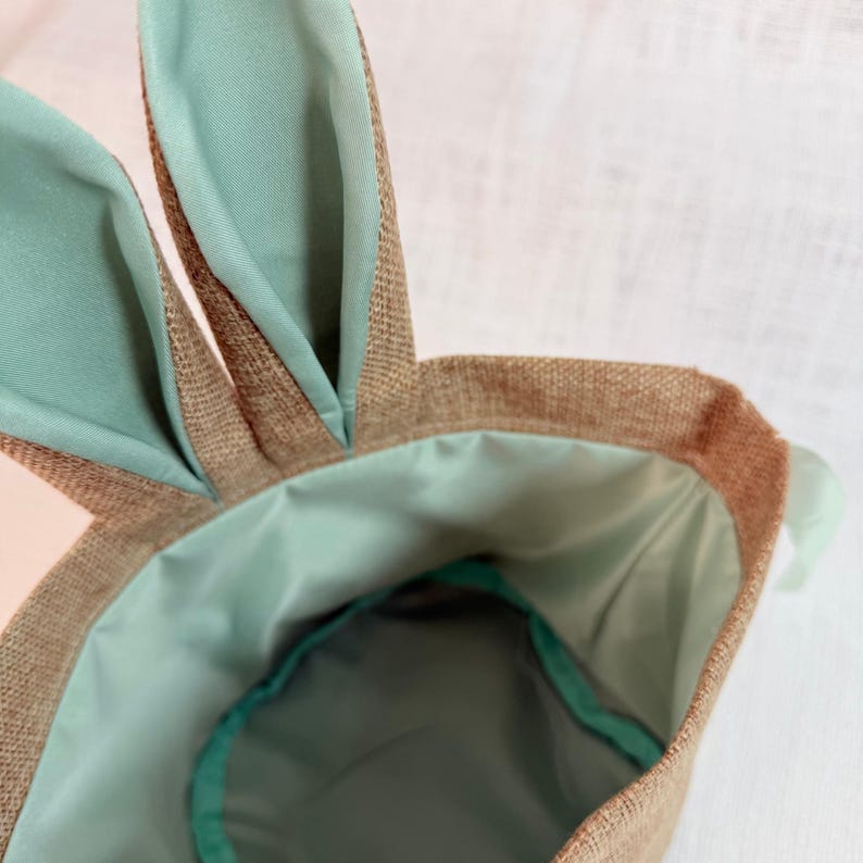 May include: A close-up of a brown burlap Easter basket with mint green accents. The basket has two bunny ears and a mint green interior. The ears and interior are made of a smooth fabric.