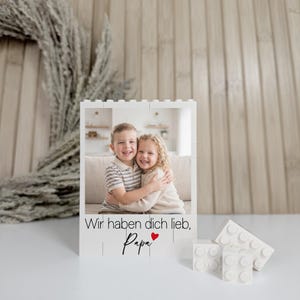 Könnte beinhalten: Ein weißer Block-Fotorahmen mit einem Foto von zwei lächelnden Kindern, die sich umarmen. Der Rahmen trägt den Text "Wir haben dich lieb, Papa" in schwarzer Schrift mit einem roten Herz. Mehrere weiße Blöcke liegen neben dem Rahmen.